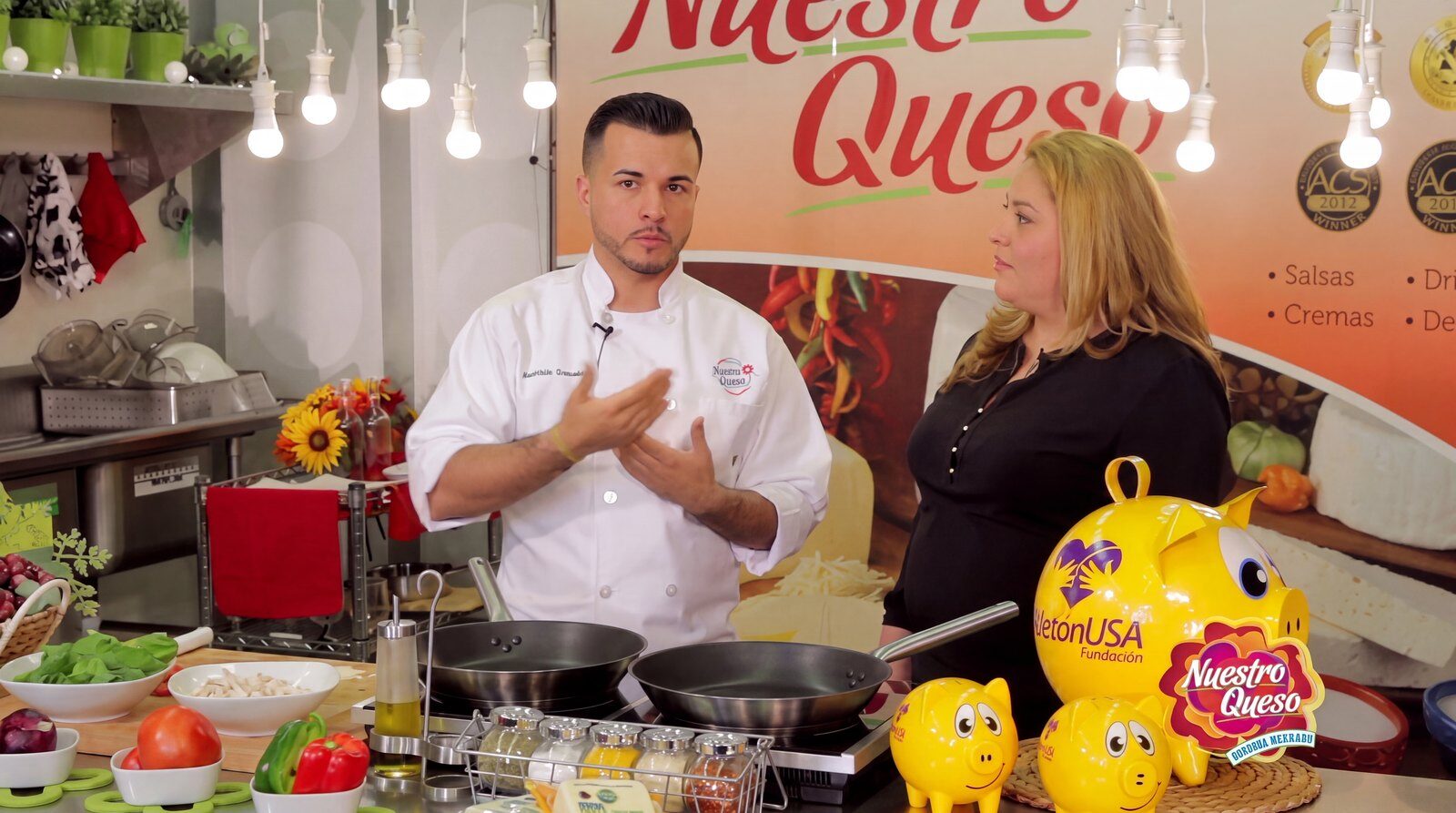 Chef Ambrocio Gonzalez in his chef’s coat, filming a cooking segment beside a presenter, with pans on the cooktop in front of him.