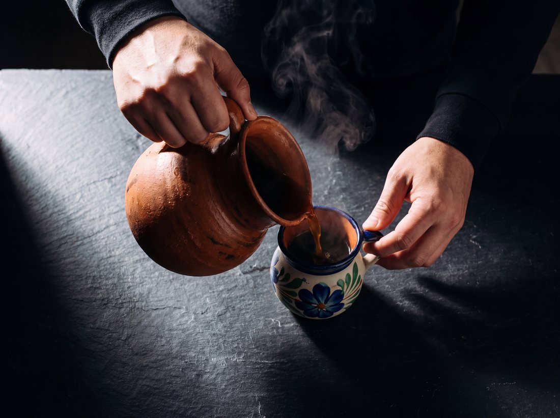 Hands pouring cinnamon coffee from a clay pitcher into a small painted clay mug.