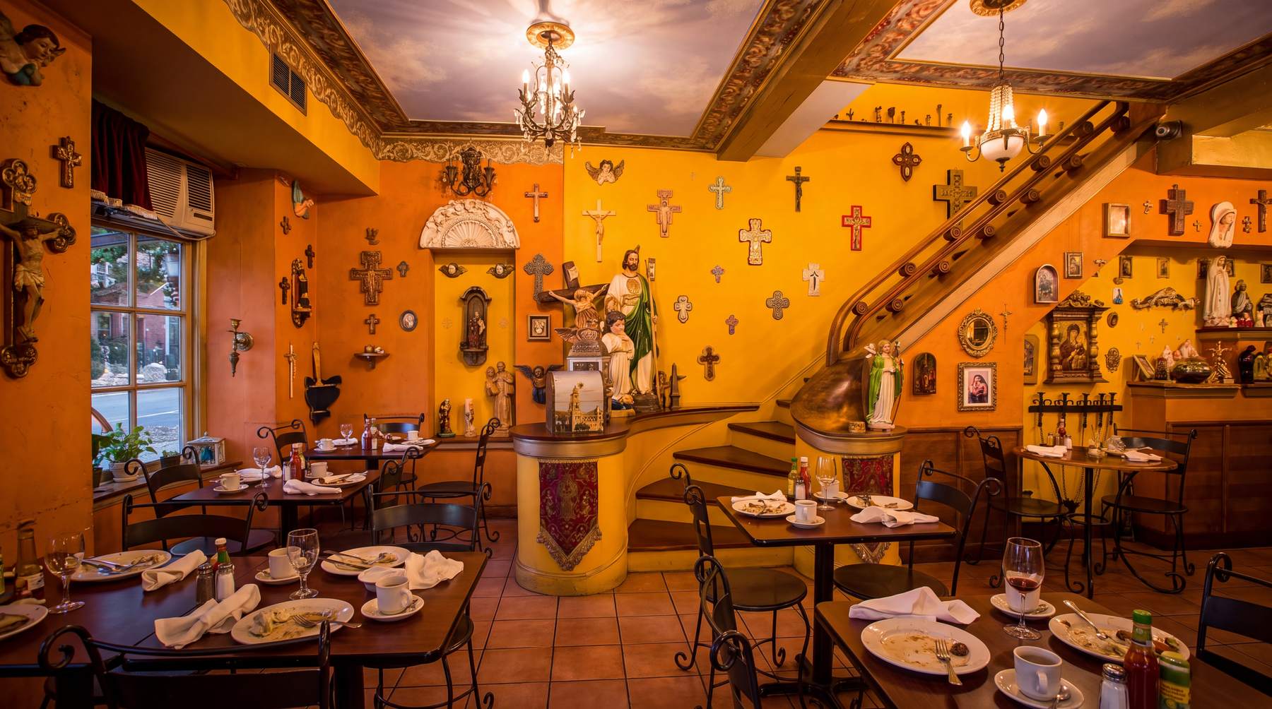 The Little Village dining room at golden hour: marigold walls covered in crosses and religious figures, a chandelier, and a painted stairway to the second floor.
