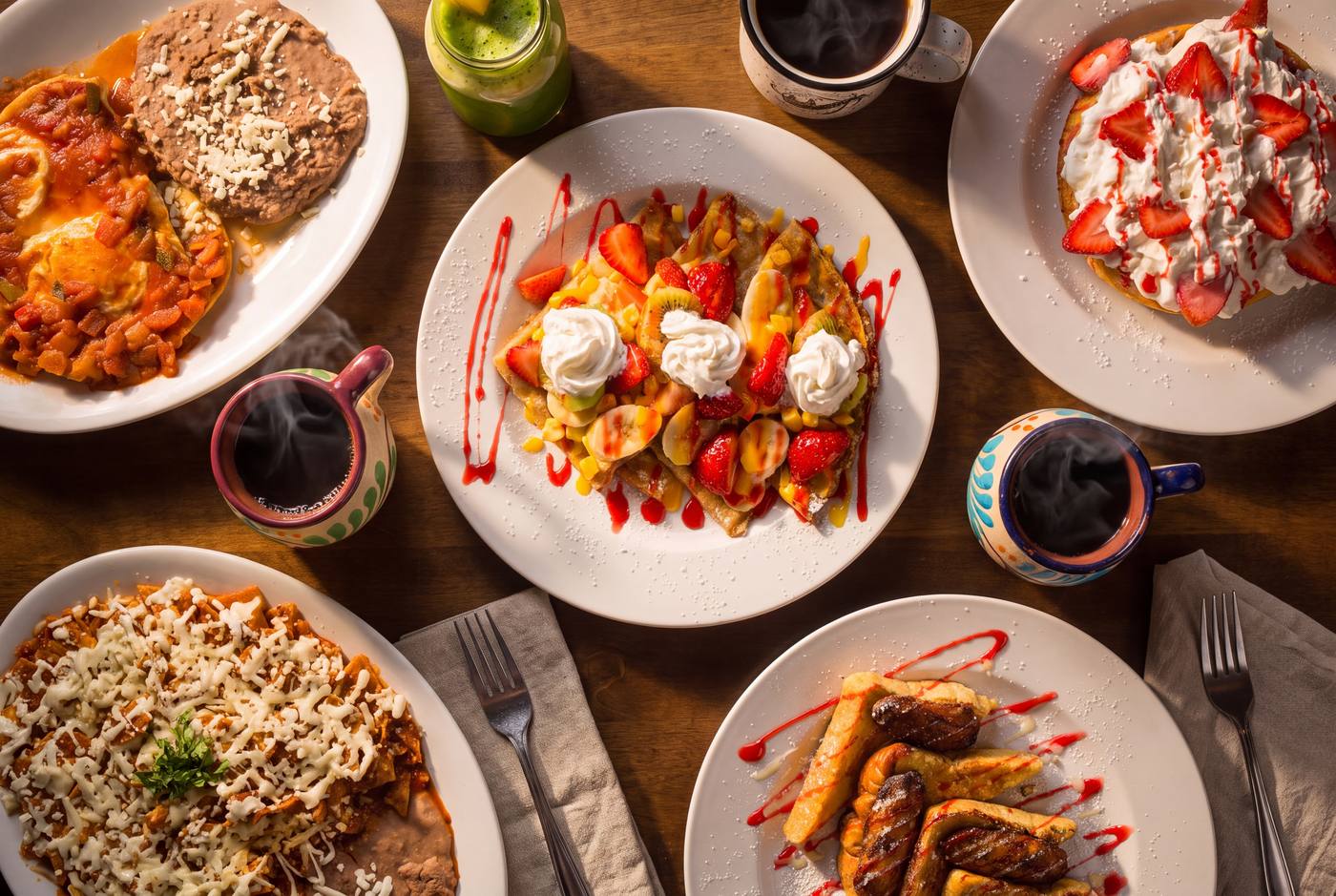 A table of Mexican breakfast dishes seen from above: chilaquiles, crepes with strawberries, coffee in painted clay mugs and a green juice.